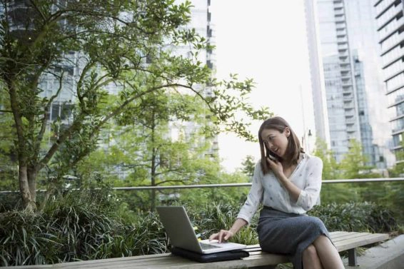 Businesswoman talking on cell phone and using laptop on bench in city