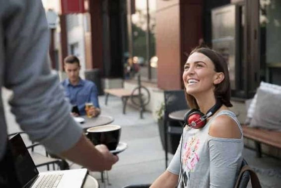 Waiter serving coffee to smiling woman using laptop at sidewalk cafe