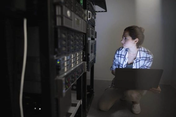Female technician with laptop working at server panel in dark server room
