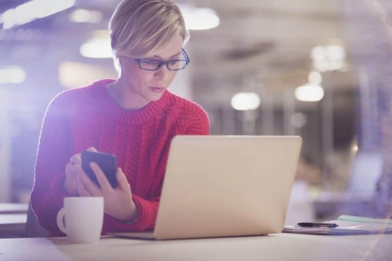 Serious businesswoman working late, using cell phone at laptop in office