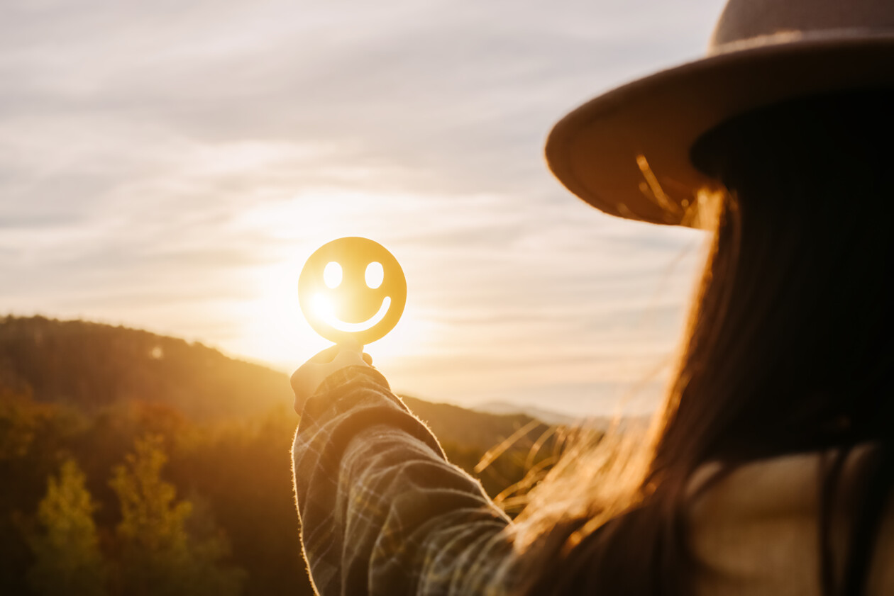 Close up of young woman holding happy smile face on background epic sunset in mountains at autumn sunny day, good feedback rating and positive customer review, satisfaction survey, mental health