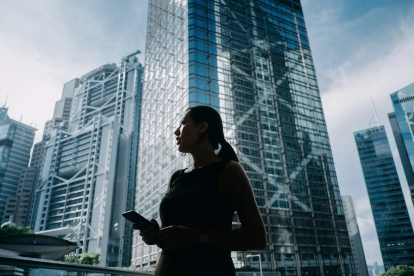 Silhouette of professional young businesswoman holding smartphone while standing against contemporary financial skyscrapers and looking up into sky with positive emotion