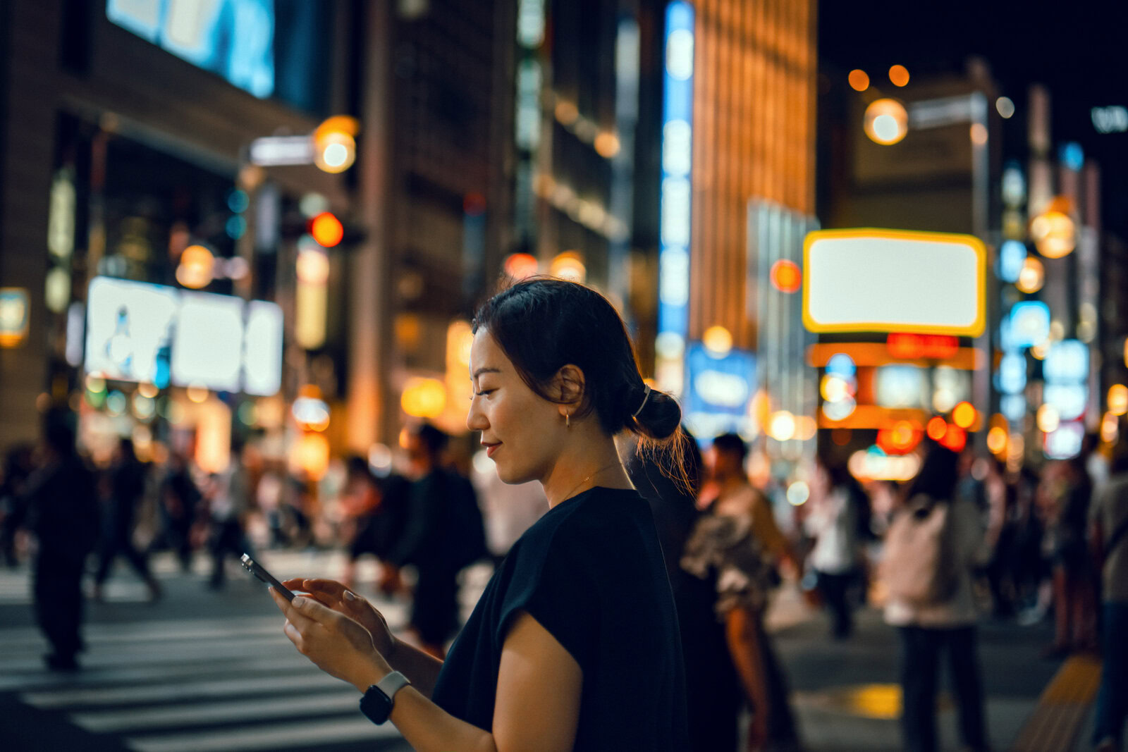Beautiful young asian woman using smartphone while commuting in the city at night, walking against busy downtown city street with multi coloured and illuminated skyscrapers and crowd of pedestrians. lifestyle and technology