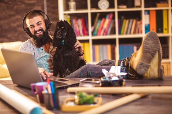 Handsome tattooed man working at home on laptop while sitting at the  table with cute dog