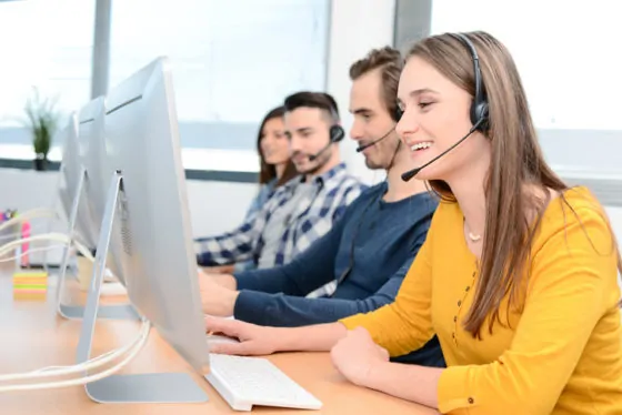 Portrait of a beautiful and cheerful young woman telephone operator with headset working on desktop computer in row in a customer service call support helpline business center with teamworker in background