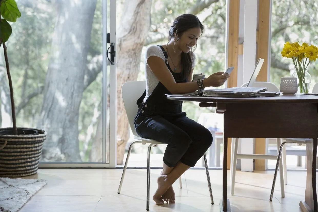 Woman texting with cell phone at laptop at dining table