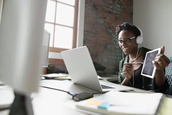 Smiling creative businesswoman with headphones holding digital tablet and video conferencing at laptop in office