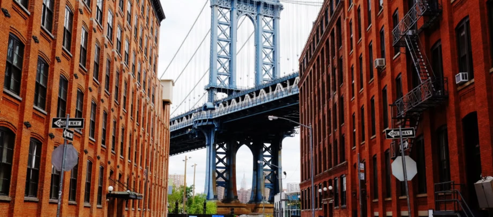 Manhattan Bridge tower framed between two red-brick buildings on a Brooklyn street, with one-way signs and fire escapes.